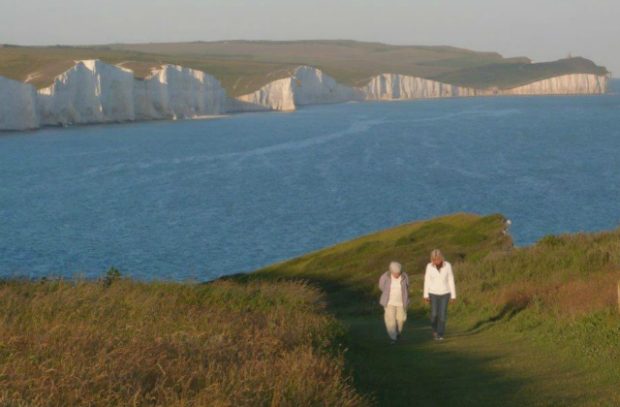 Two people walking on a coastal path against a backdrop of white cliffs and the sea.
