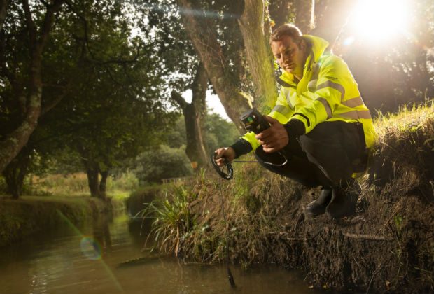 An image of a man kneeling by a river wearing a high vis yellow jacket.