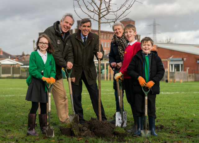 An image of Minister Rutley standing in a field planting a tree with Tree Champion Sir William Worsley and pupils from St Andrew’s CE Primary School. They are planting a tree.