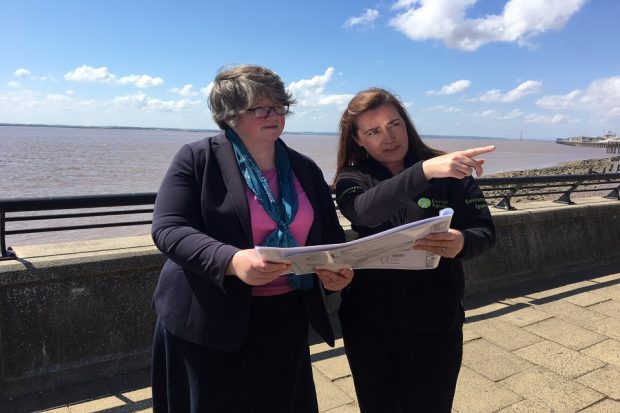 Image of Environment Minister Dr Thérèse Coffey being shown what part of the Humber: Hull Frontage Flood Defence Improvements will look like by scheme project manager Helen Tattersdale. They are looking at a piece of paper against a sea view backdrop.