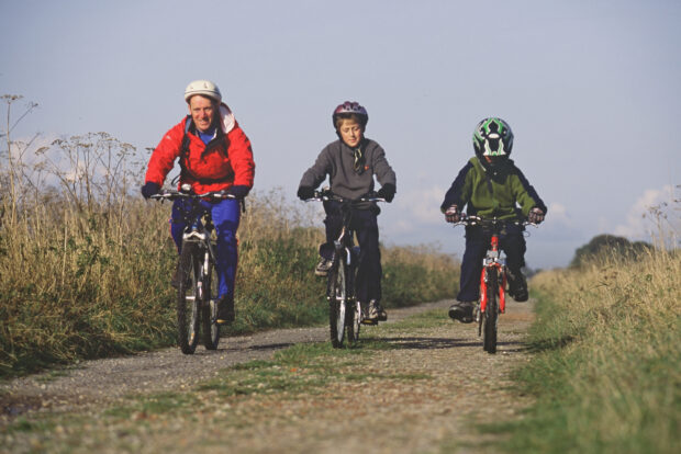 A man and two children riding bikes with helmets on down a path.