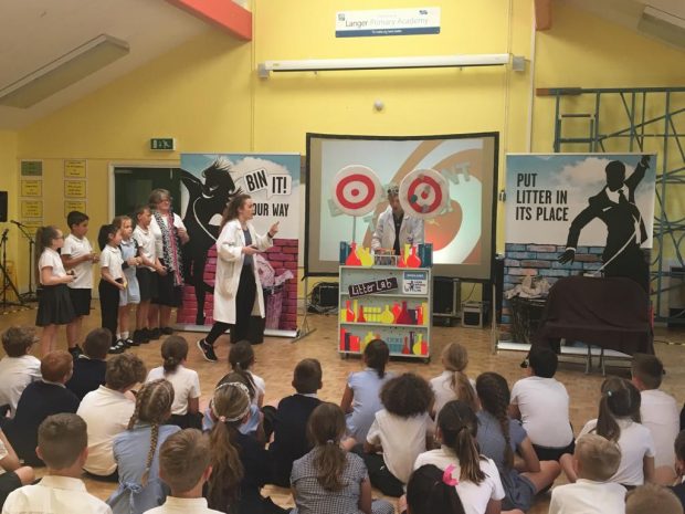 Image of school children and Minister Therese Coffey standing up in front of an audience of children at the Bin It Roadshow. The sign on the wall says ‘bin it your way’ and ‘put litter in its place’