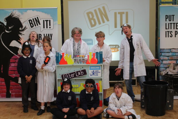 Image of Minister Therese Coffey standing with children and an actor behind a sign that says ‘Litter Lab’ and ‘Bin It!’