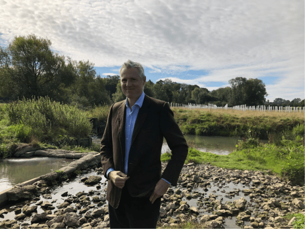 Image of Lord Goldsmith in front of a riverbank with trees in background. 