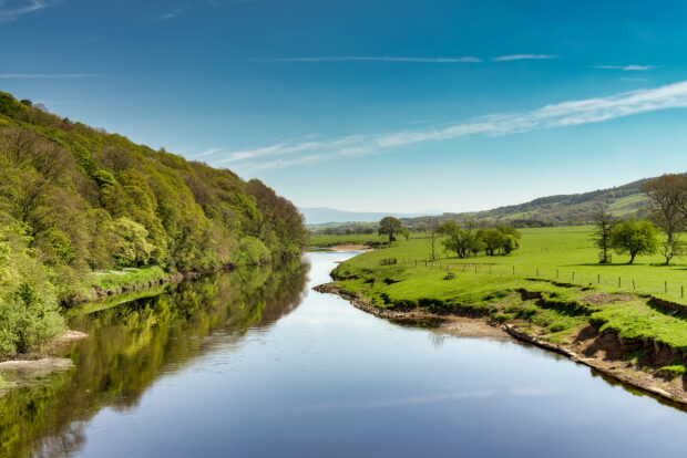 A view of the River Lune near Lancaster on a sunny day, with green fields and wooded slopes.